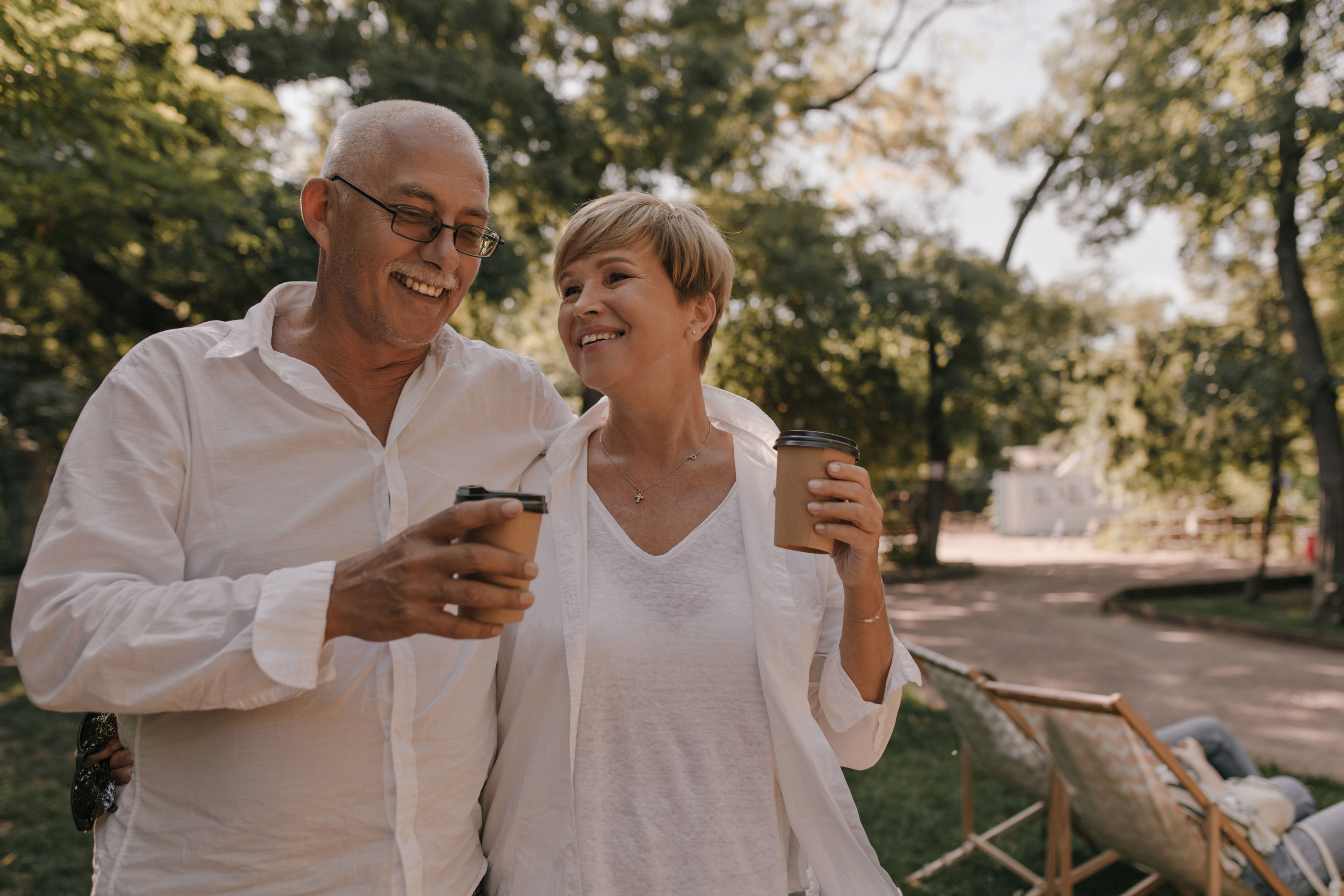 grey haired man with eyeglasses in white shirt smiling and posing with cap of tea and stylish woman in light outfit outdoor