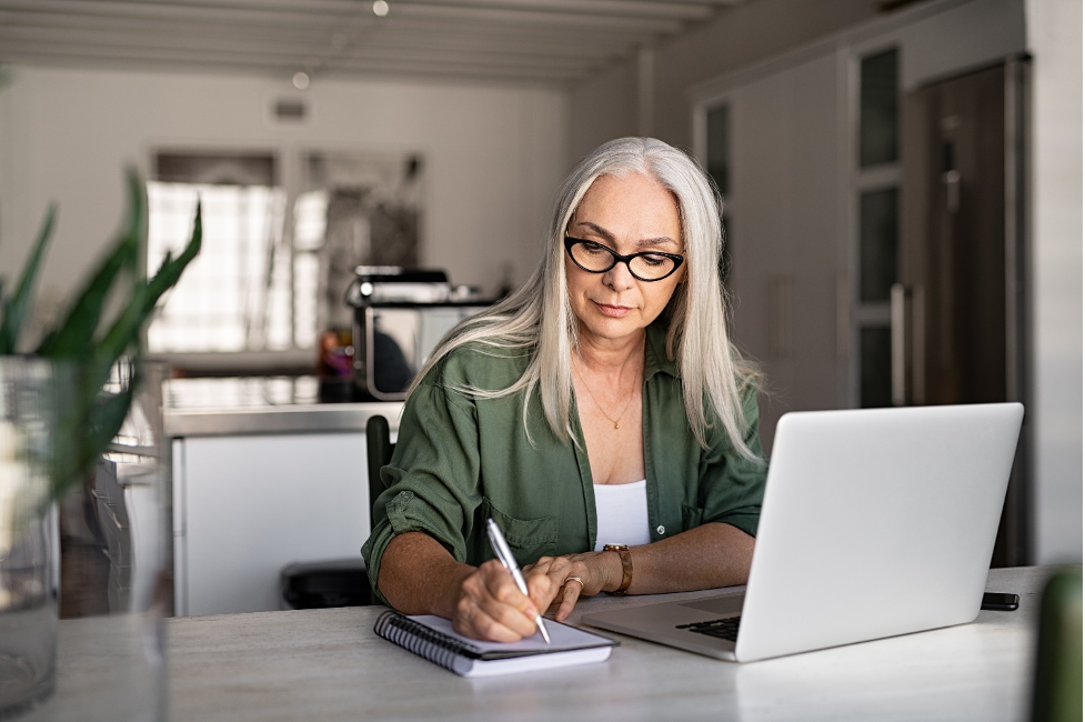 Senior stylish woman taking notes in notebook while using laptop at home. Old freelancer writing details on book while working on laptop in living room. Focused cool lady writing notary in notepad by Ridofranz/istockphoto