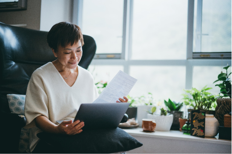 Senior Asian woman looks over financial documents with her laptop at home.